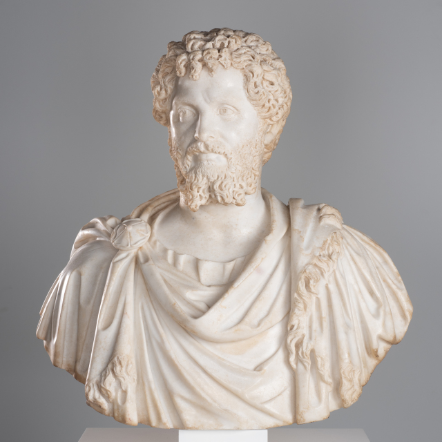 A white marble bust of a man with curly hair and beard. He glances to his left.  He wears a tunic and fringed cloak, fastened at his left shoulder with a circular brooch.