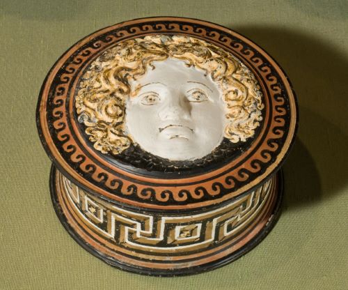 A round box is decorated with a geometric pattern.  The lid, which also has a patterned border, is decorated with a woman's face that is raised in relief.  She has white skin, curly yellow hair, and light brown eyes. 