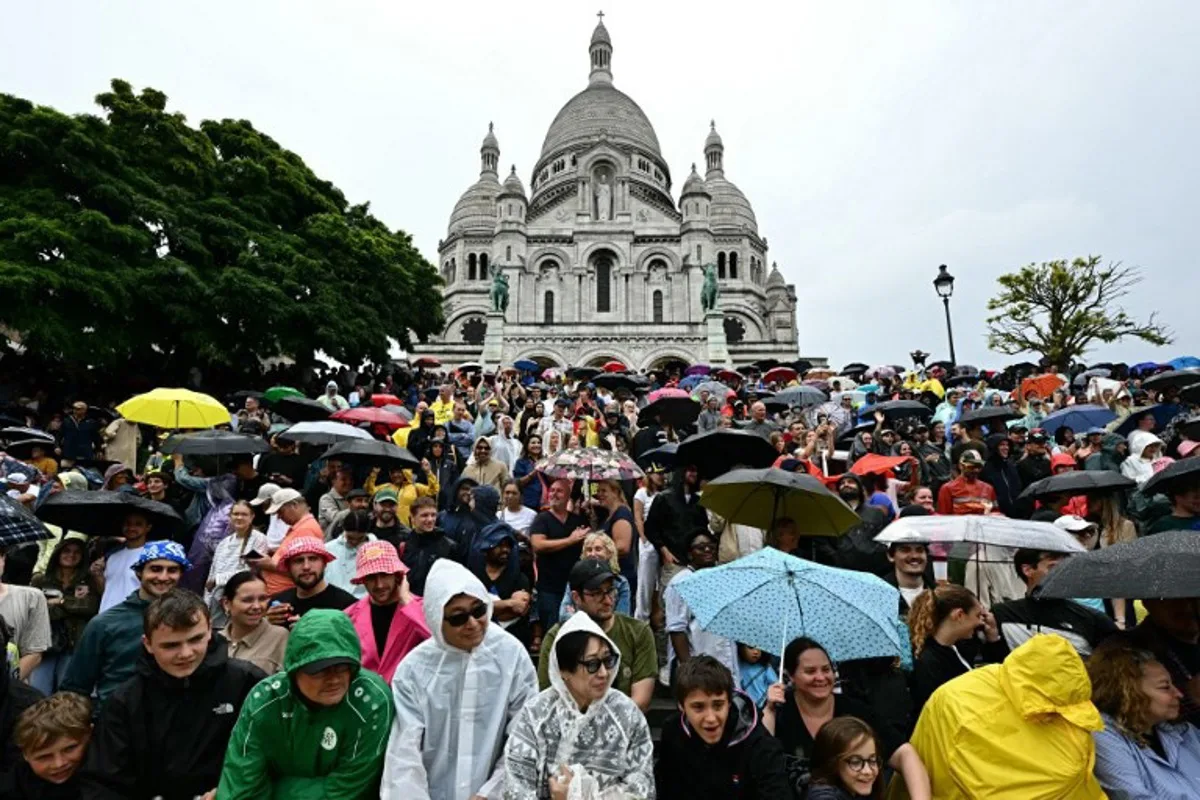Spectators await the pack of riders (peloton) in the rain, at the foot the Sacre-Coeur Basilica on the Butte de Montmartre during the 21st and final stage of the 112th edition of the Tour de France cycling race, 132.3 km between Mantes-la-Ville and Paris' Champs-Elysees Avenue, on July 27, 2025. Loic VENANCE / AFP