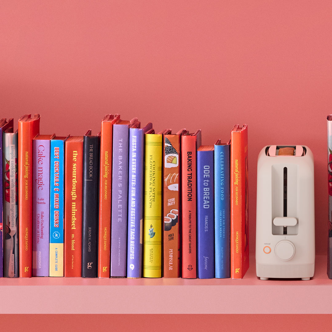 The bella® 2-Slice Slim Toaster in Oatmilk sits neatly on a vibrant pink shelf beside a row of colorful cookbooks — a compact, stylish statement piece made to fit anywhere.
