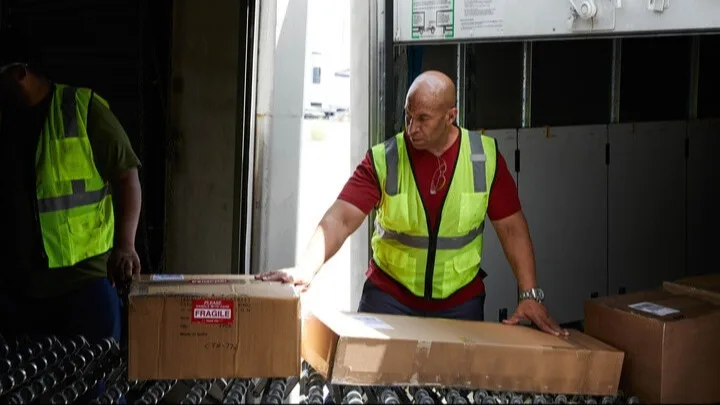Warehouse workers in safety vests handling cardboard boxes marked "fragile" near a loading dock and roller conveyor.