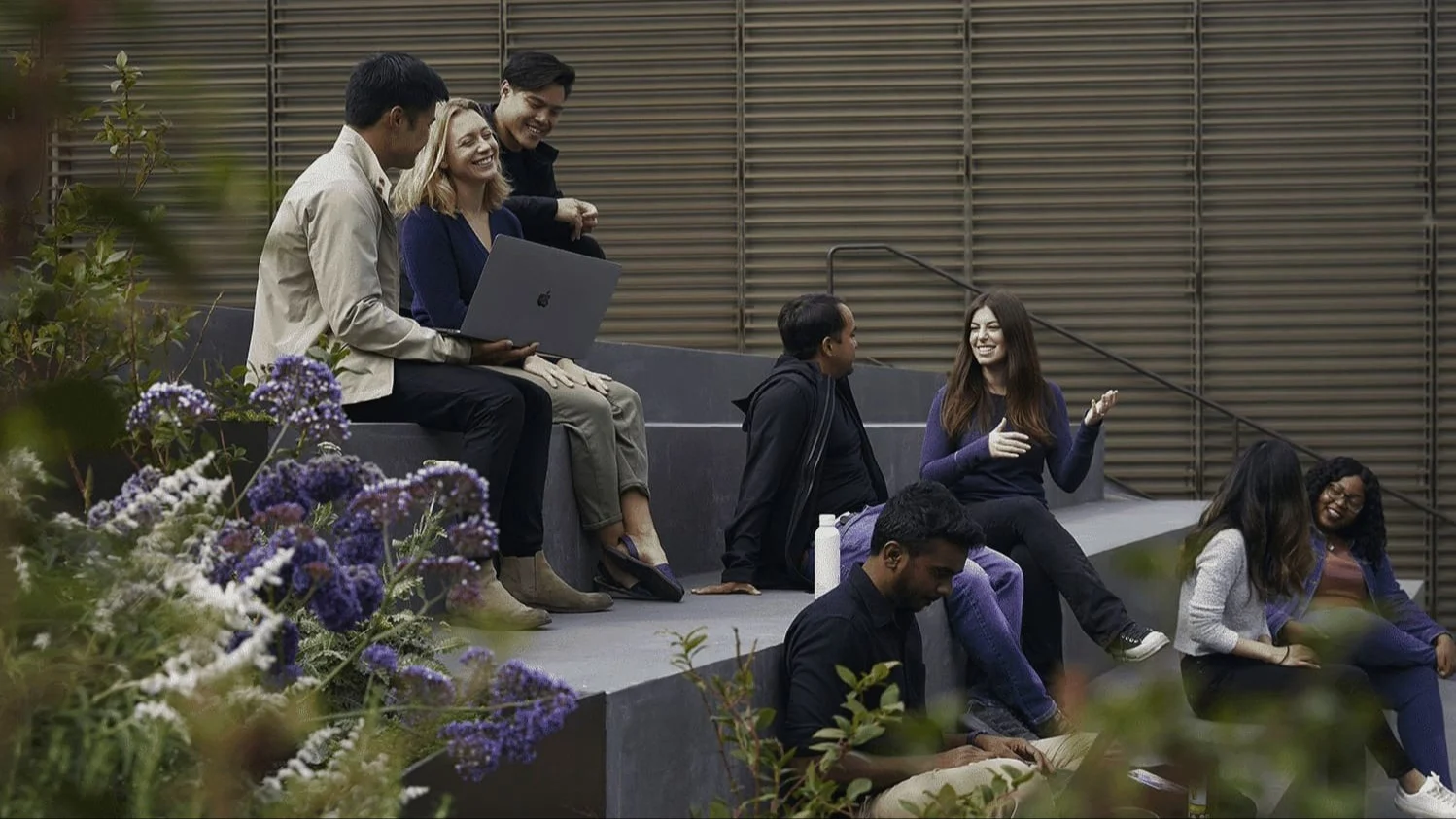 Group of diverse young professionals socializing outdoors on concrete steps with laptop, surrounded by purple flowers.