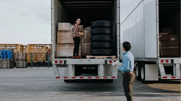 Person standing in truck loaded with boxes and tires while another person with clipboard stands outside.