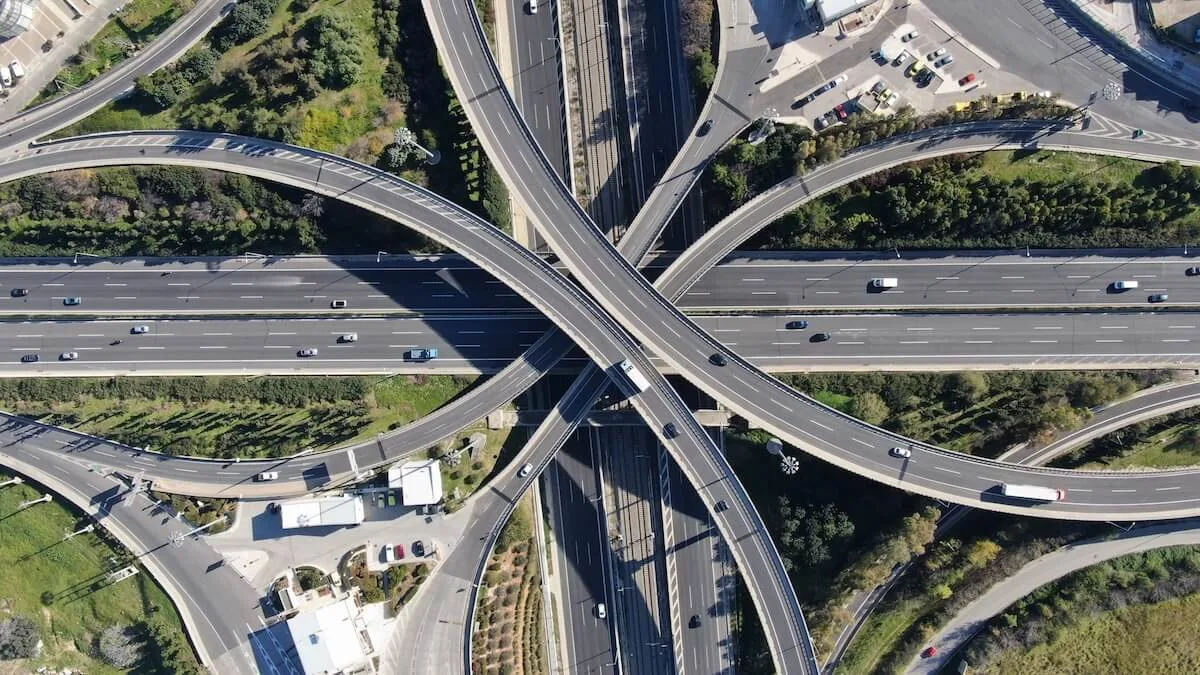 Top-down aerial view of a complex highway interchange with multi-level overpasses, curving ramps, and light vehicle traffic traveling along intersecting roadways surrounded by green trees and fields.