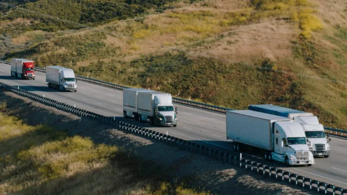 Semi trucks traveling on a highway through rolling grasslands, aerial view of freight transport convoy.