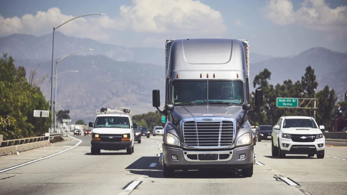 Silver semi truck driving on highway with mountains in background, surrounded by other vehicles on a sunny day.
