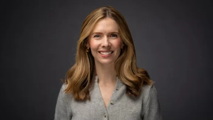 Headshot portrait of Uber Freight CEO Rebecca Tinucci smiling in a grey short-sleeved knit top against a dark studio background.
