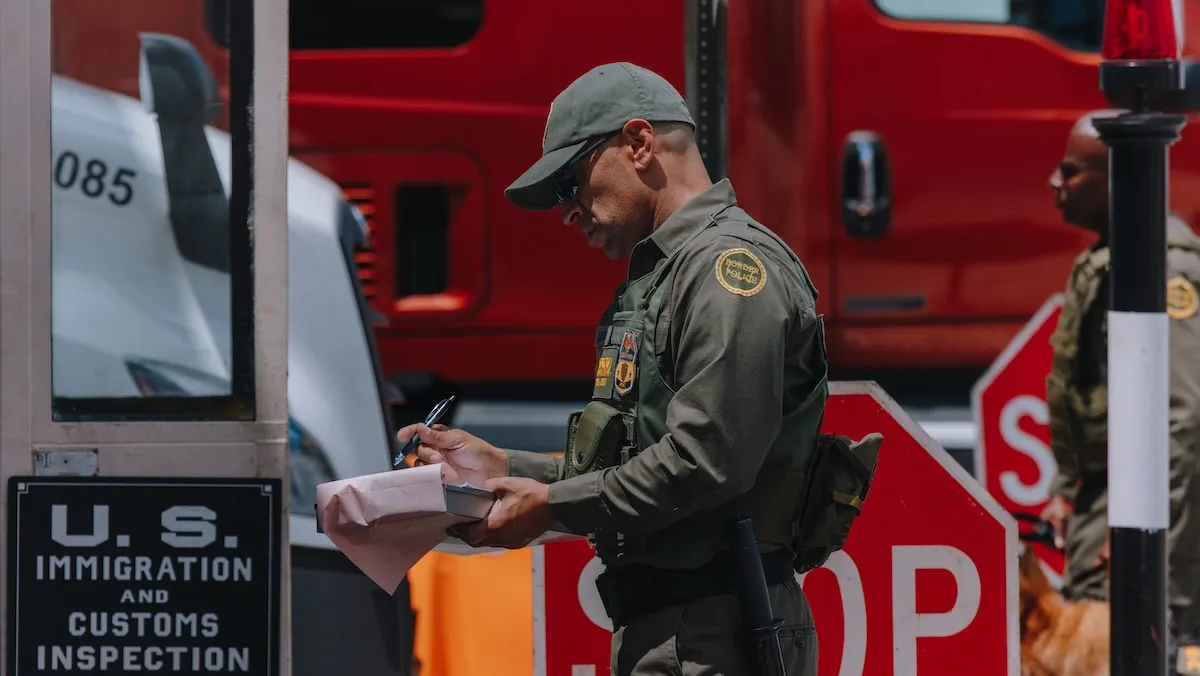 A U.S. customs officer in a green uniform reviews freight paperwork on a clipboard at a border inspection checkpoint. A red commercial truck and another officer with a K9 are in the background.