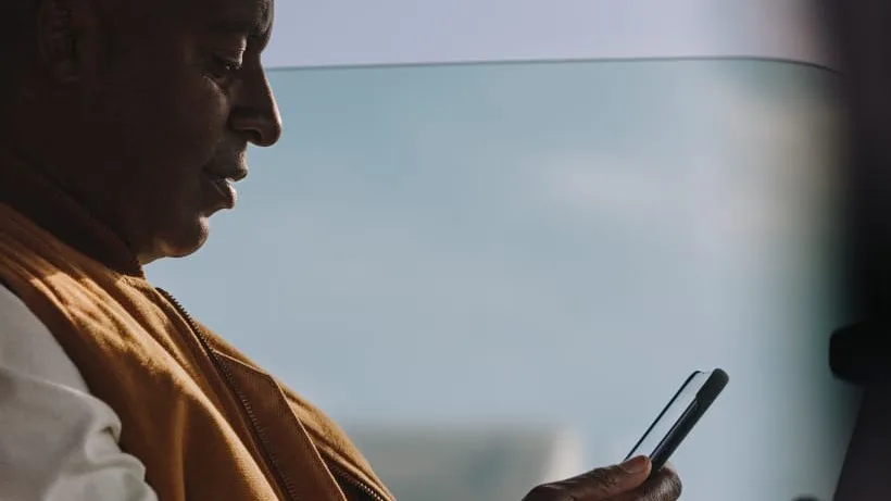 Person in brown jacket looking at smartphone while traveling, silhouetted against bright window light.
