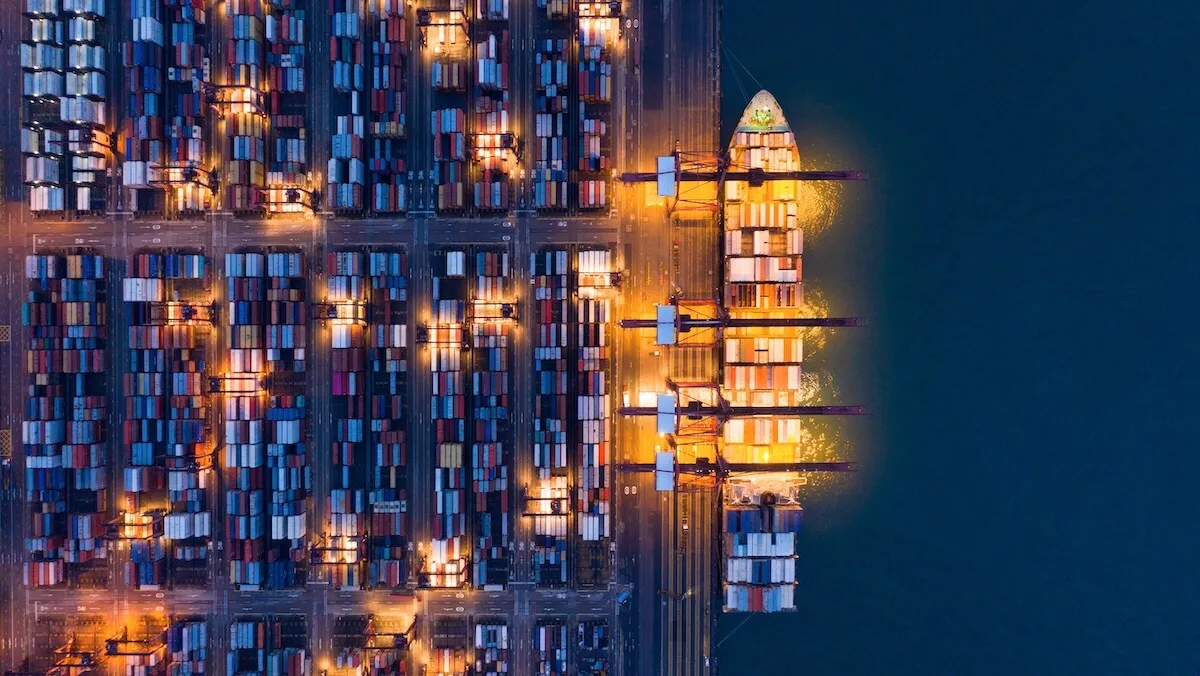 Aerial, top-down night view of a bustling container port with a cargo ship docked on the right, illuminated gantry cranes, and grid-like stacks of colorful shipping containers.