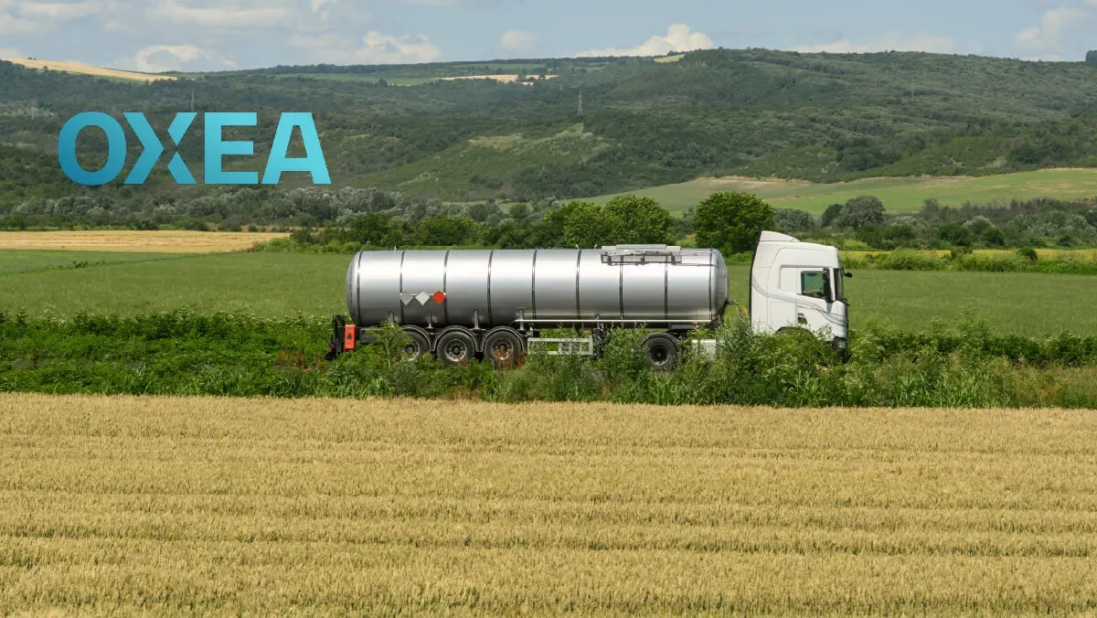 A white semi-truck pulling a silver cylindrical tanker trailer drives along a rural road, framed by a golden field in the foreground and green rolling hills in the background, with a light blue "OXEA" logo overlaid in the top left corner.