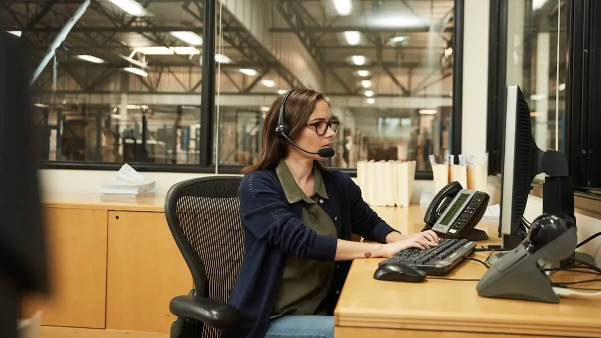 Une spécialiste en logistique portant un casque et des lunettes, assise à un bureau en train de taper sur un ordinateur de bureau, avec de grandes fenêtres derrière elle donnant sur un entrepôt.