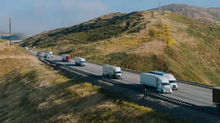Semi trucks traveling on a highway winding through rolling hills under clear blue sky