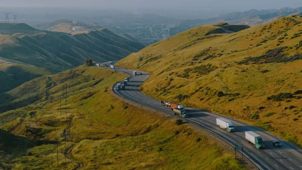 Route sinueuse avec camions et voitures traversant des collines dorées, avec montagnes et ville visibles dans la brume au loin.