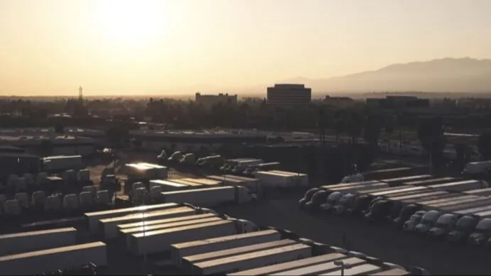 Sunset over a truck depot with semi-trailers parked in rows, city buildings and mountains silhouetted in the background.