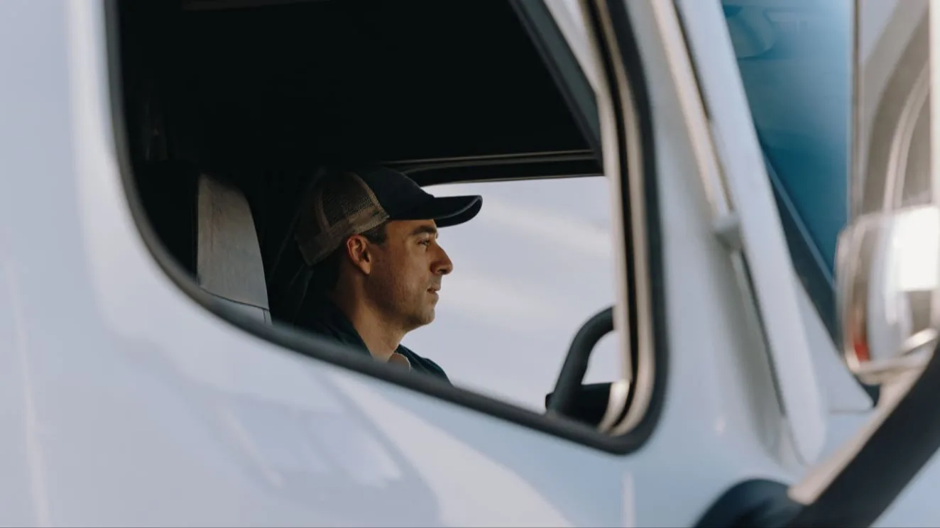 Side view of a truck driver wearing a trucker cap visible through the window of a white delivery vehicle.