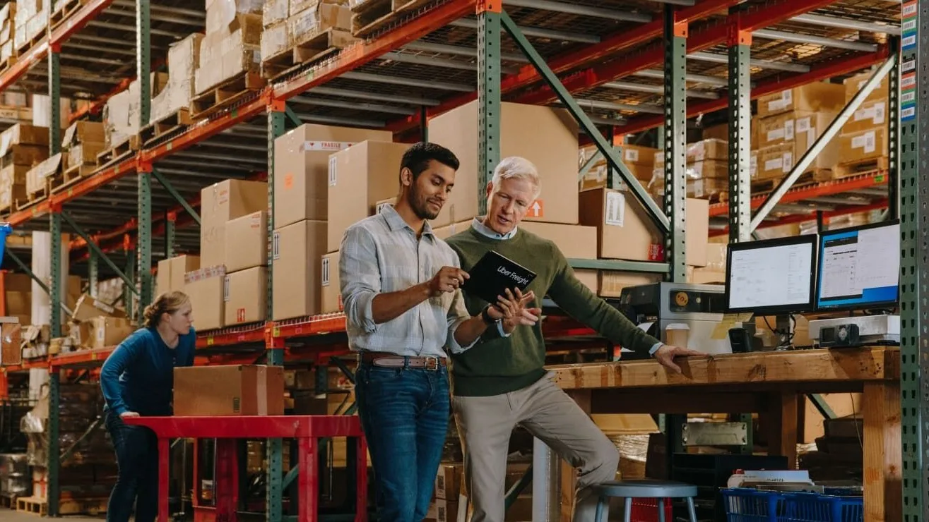 Two colleagues reviewing documents in a warehouse with tall shelving racks filled with cardboard boxes and inventory.