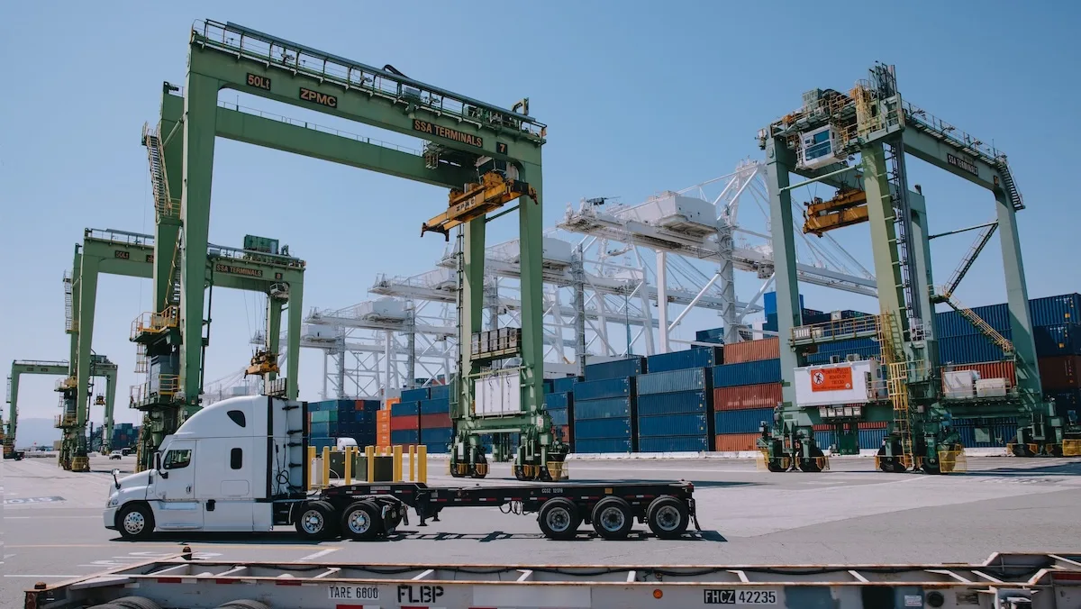 Un semi-remorque blanc dans un port maritime, entouré de grandes grues portiques vertes et de conteneurs de fret colorés empilés.