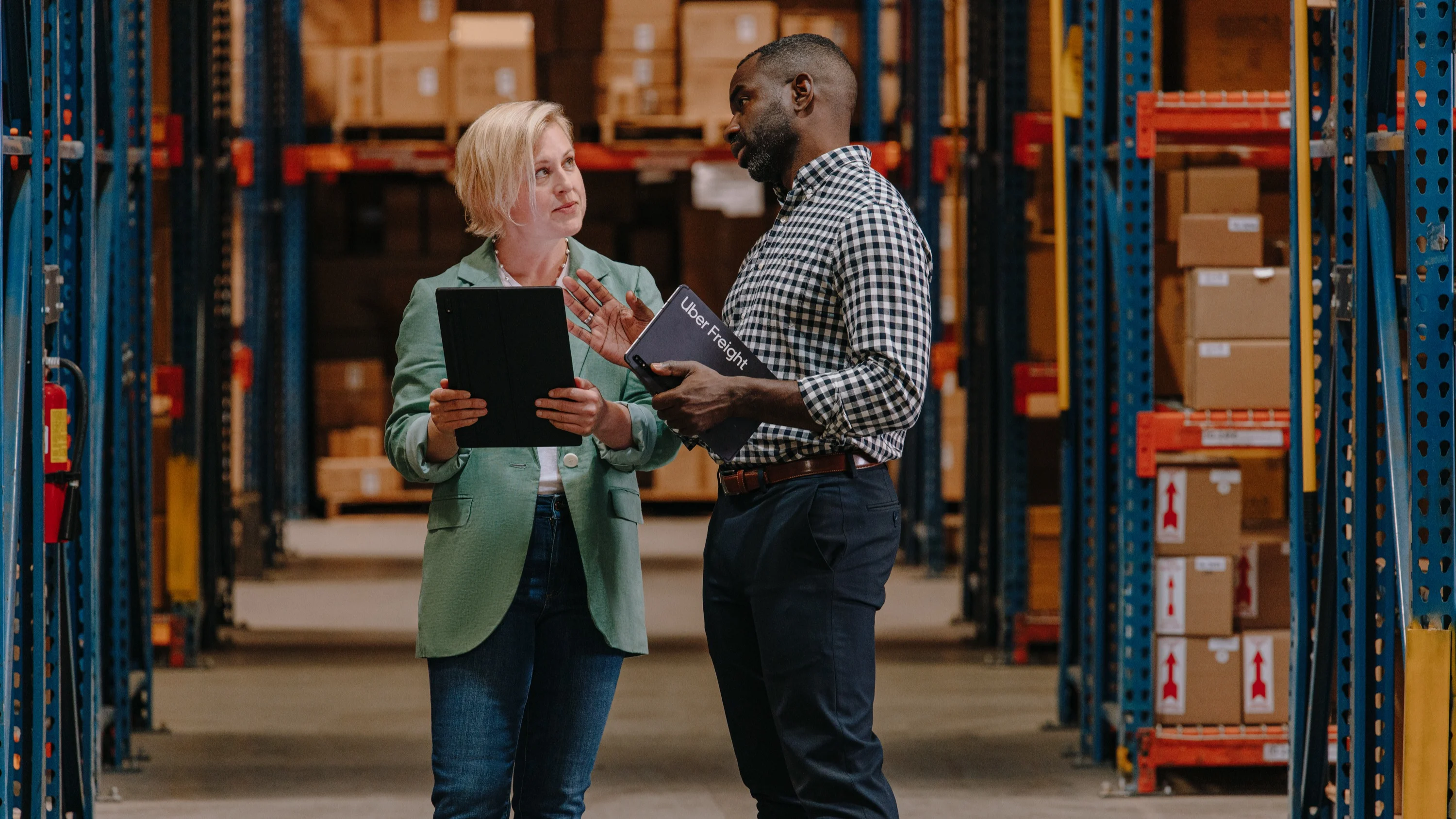 Two colleagues discussing inventory in a warehouse, one holding a tablet while the other reviews documents.