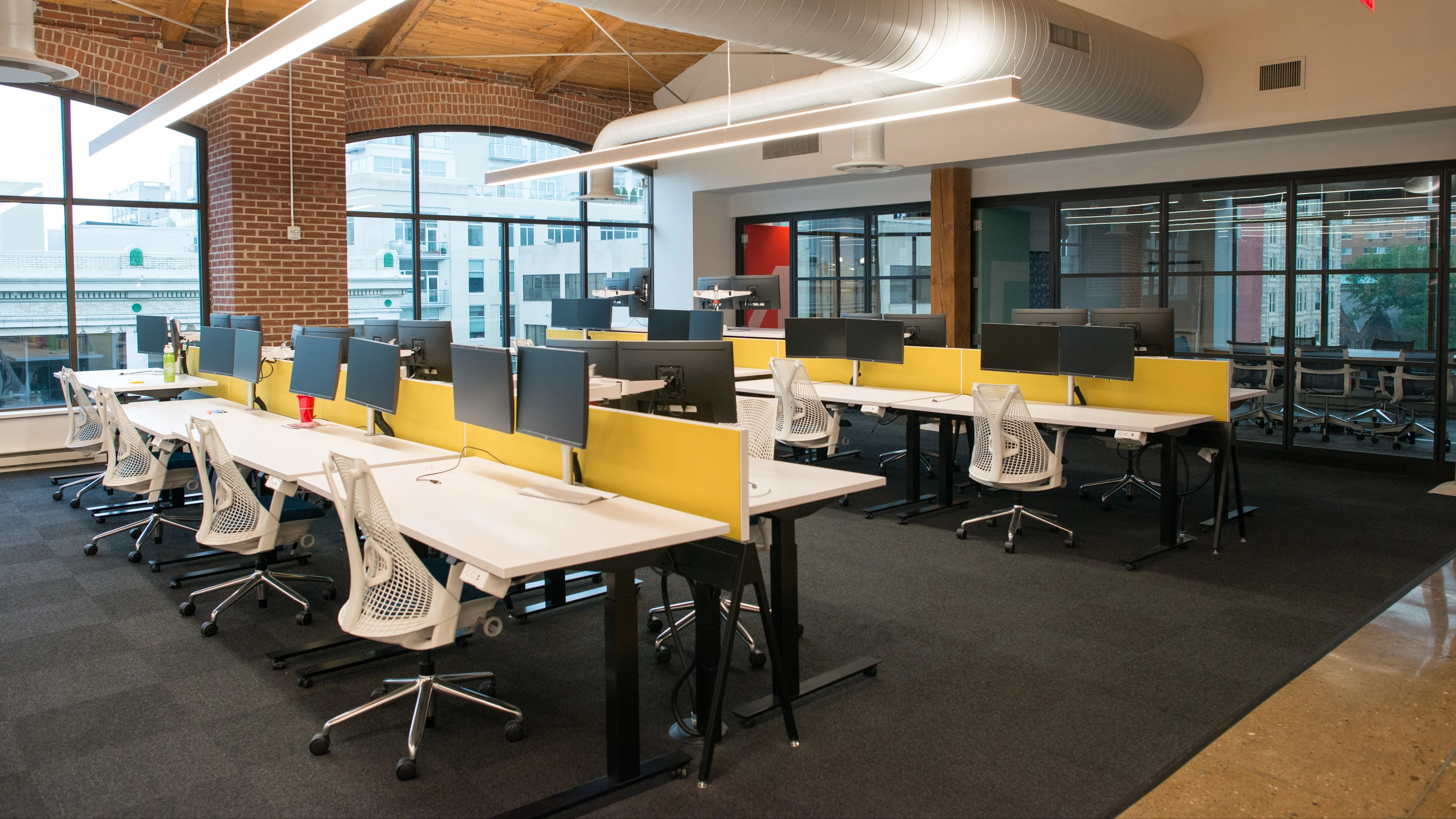 Espacio de coworking moderno con escritorios blancos, divisores amarillos y pantallas de privacidad azules en una oficina de paredes de ladrillo con vistas a la ciudad.