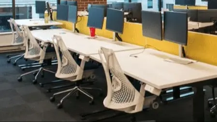 Modern coworking space with white desks, yellow dividers, and blue privacy screens in a brick-walled office with city views.