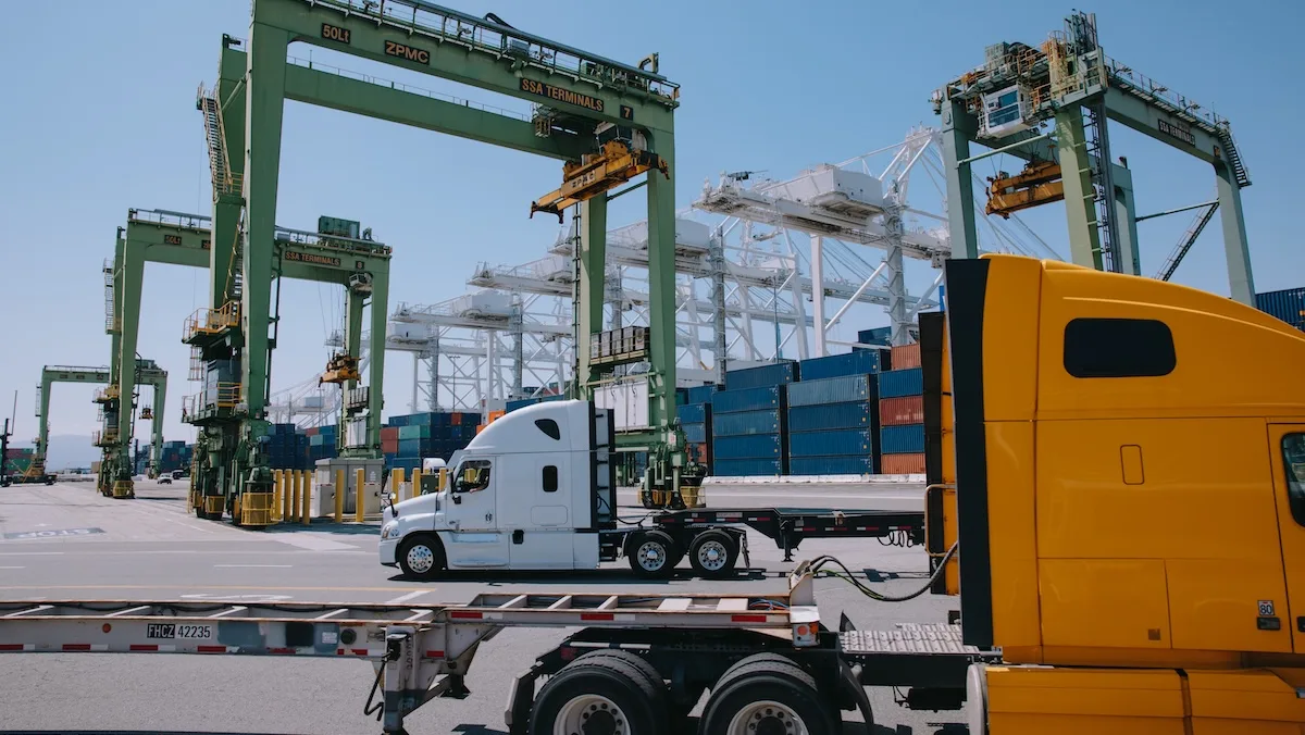 Semi trucks and cargo containers lined up at a busy shipping port, with large green container cranes towering overhead against a clear sky.