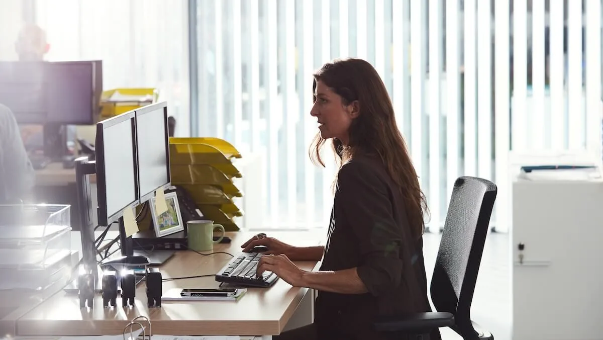 Logistics office worker sitting at a desk, focused on a dual‑monitor computer setup in a bright, modern office with vertical window blinds in the background.