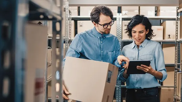 Two colleagues in denim shirts checking inventory on a tablet while managing boxes in a warehouse storage area.