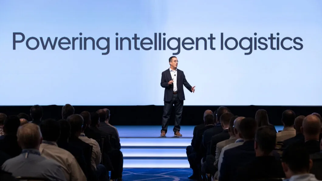 Speaker presenting on stage at a conference with "Powering intelligent logistics" displayed on the blue backdrop screen.