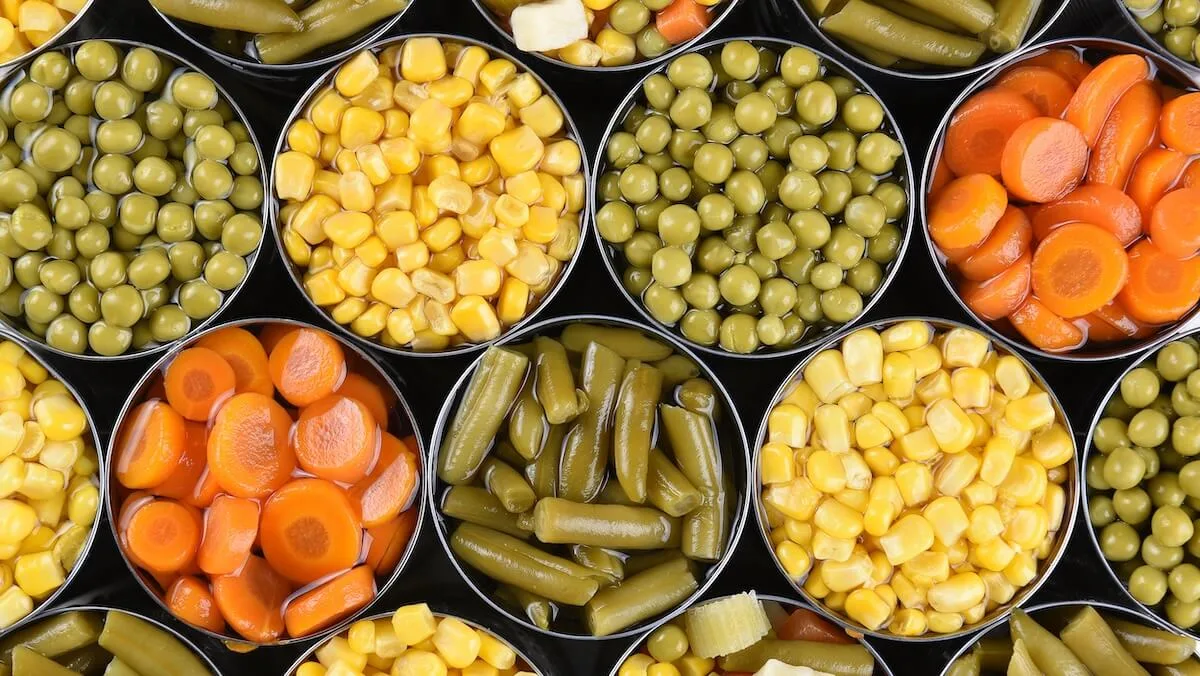 Top-down view of open cans filled with mixed vegetables—green beans, peas, corn, and sliced carrots—arranged tightly in a colorful grid pattern.