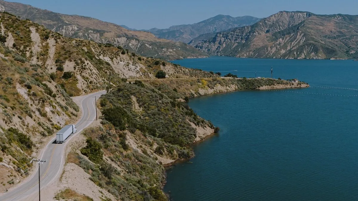 Camion sur une route à voie unique traversant des montagnes et longeant un plan d'eau.