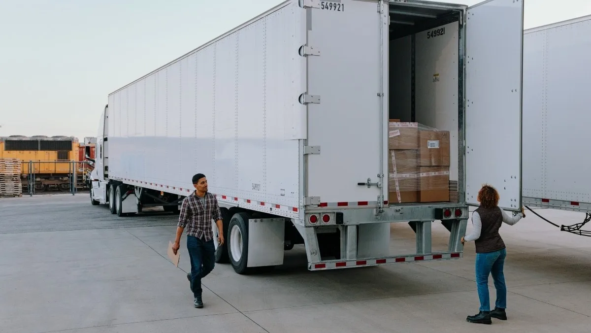 Line of semi‑trucks and cars driving along a divided highway that winds through green, hilly terrain under a clear sky.