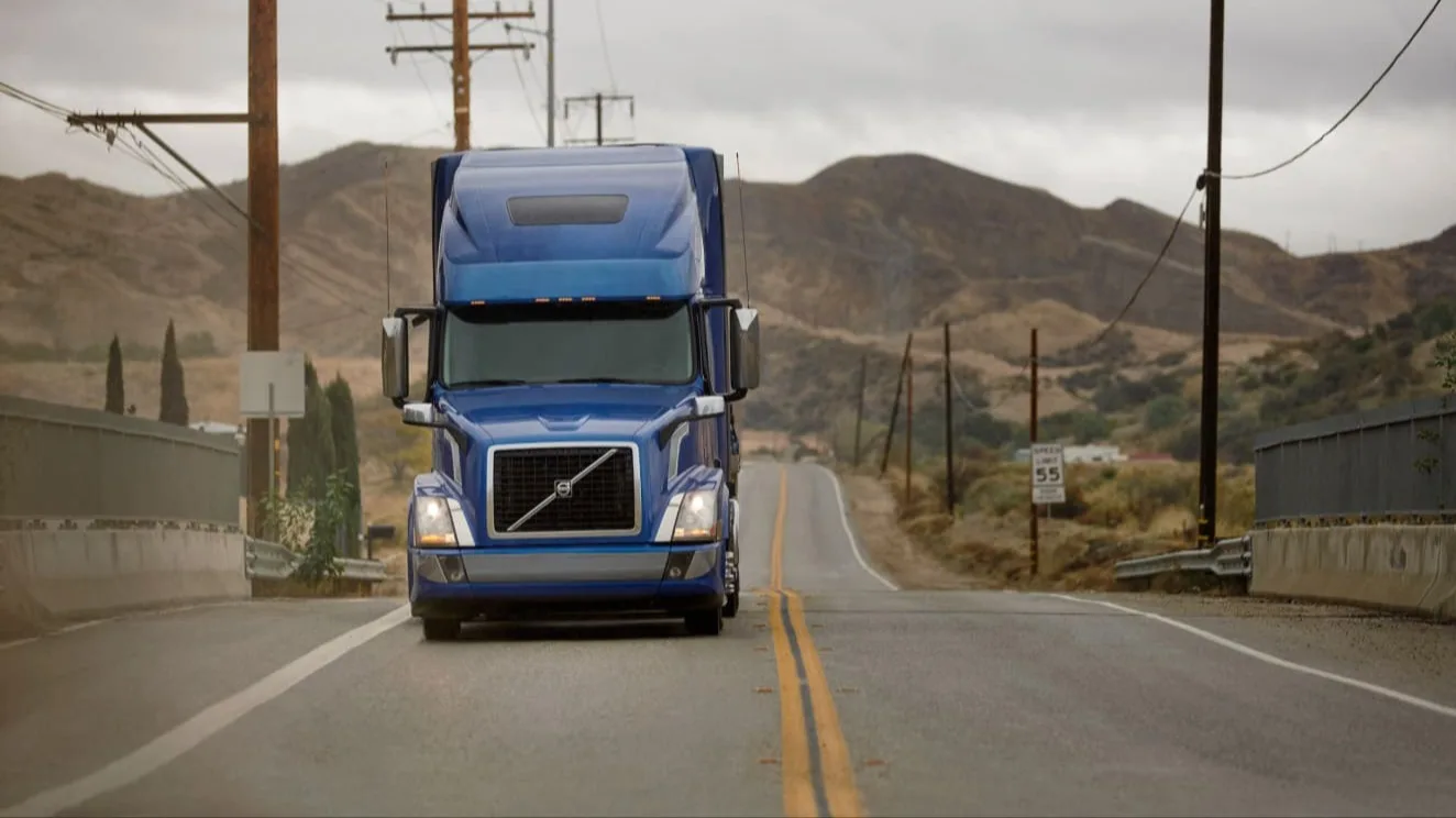 Blue Volvo semi-truck driving on a desert highway with mountains in background under overcast skies.