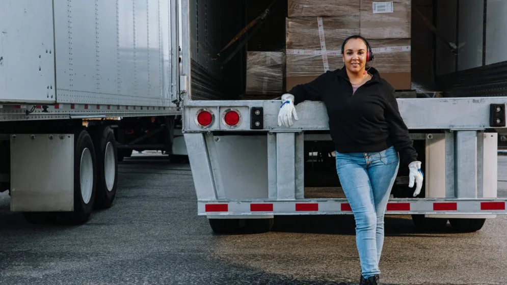 Worker in black jacket and white gloves standing beside open delivery truck loaded with wrapped packages.