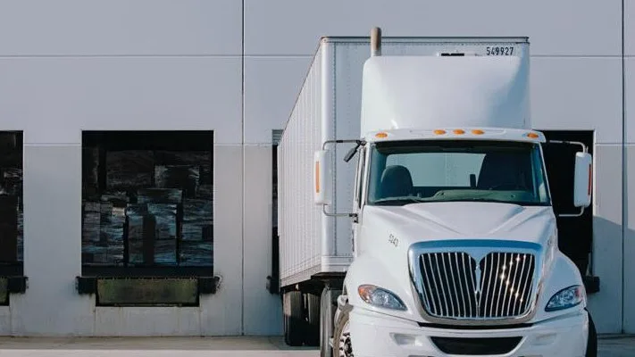 White delivery truck parked at loading dock with two bay doors visible against a gray warehouse wall.