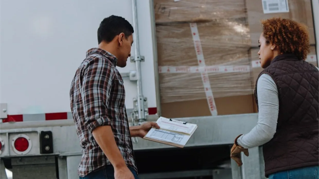 Two people examining delivery paperwork beside a truck with wrapped packages visible in the cargo area.