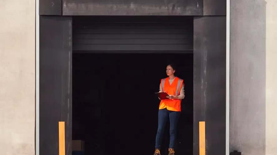 Person in bright orange safety vest holding clipboard standing in large industrial doorway with concrete walls.