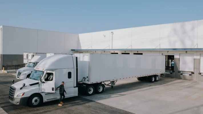 White semi truck with trailer parked at loading dock of industrial warehouse facility under clear blue sky