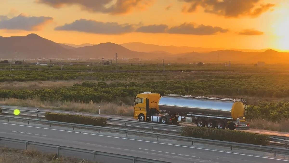 A yellow European chemical tanker truck pulling a silver metallic liquid container trailer drives along a highway through countryside fields.
