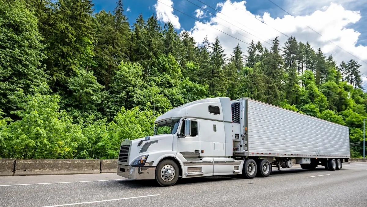 An unmarked white semi-truck pulling a long trailer drives along a paved highway with a dense green forest in the background under a partly cloudy blue sky.
