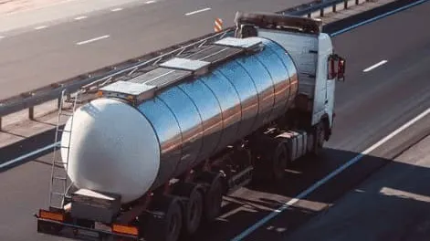 Silver tanker truck driving on highway with autumn orange trees in background during sunset.
