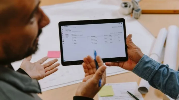 Two people reviewing data on a tablet during a business meeting with blueprints and office supplies on the table.