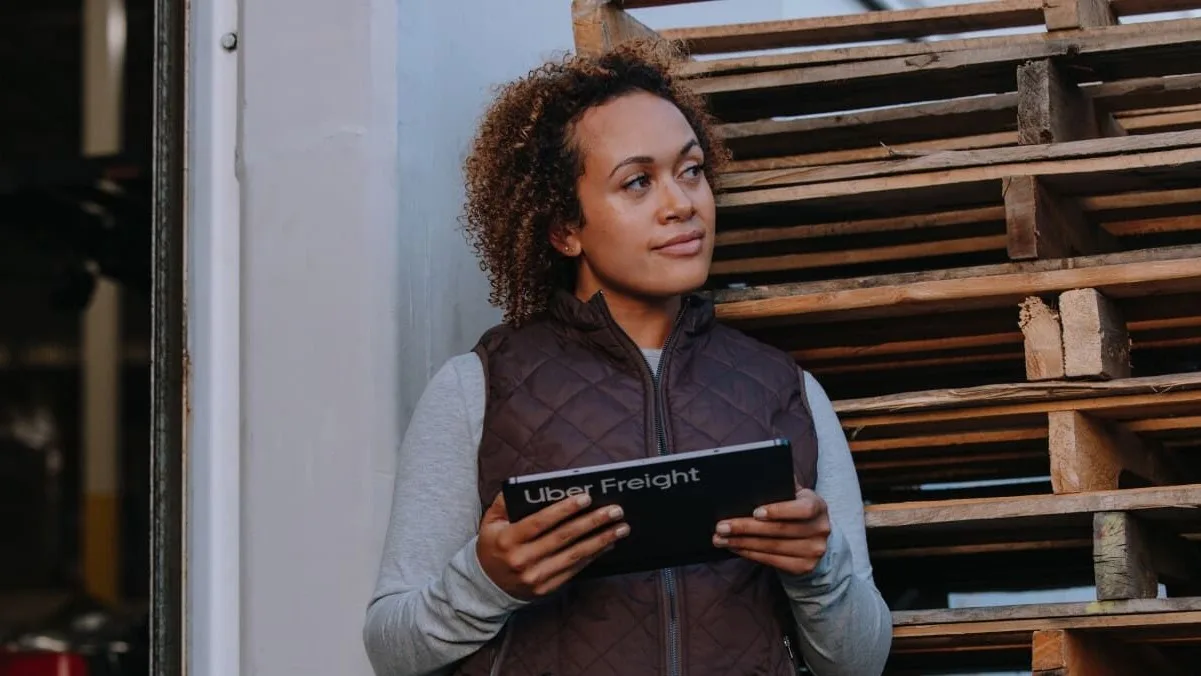 Person in brown vest holding Uber Freight tablet near wooden pallets in warehouse setting