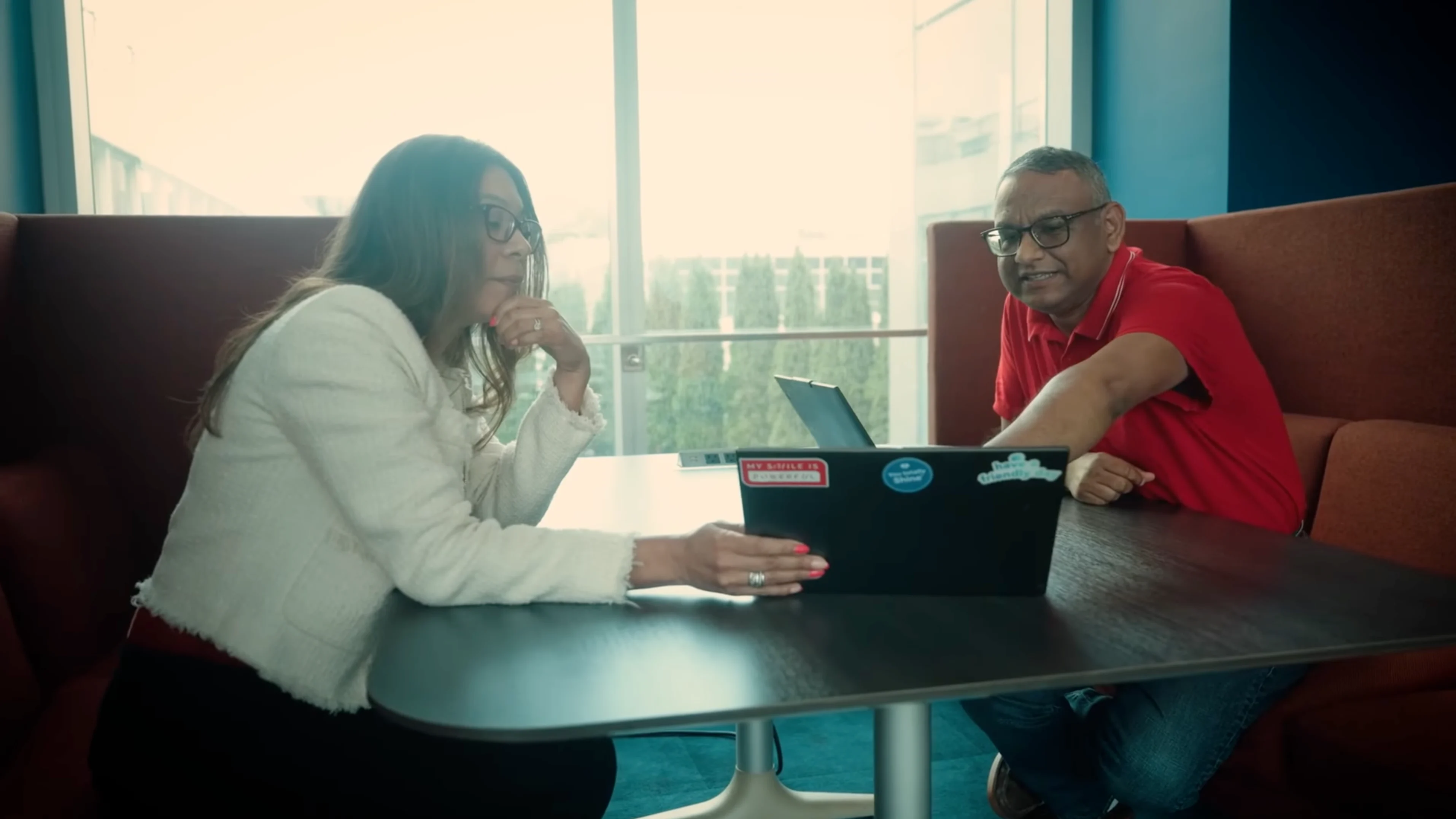 Person with long dark hair wearing red-framed glasses and light blazer, smiling while speaking in an indoor setting.