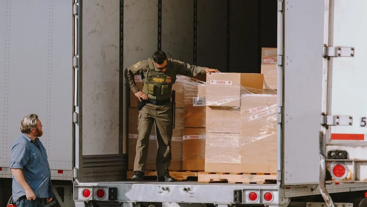 A US Border Patrol agent inspecting shrink-wrapped palletized cargo boxes inside the back of a freight trailer at a cross-border checkpoint.