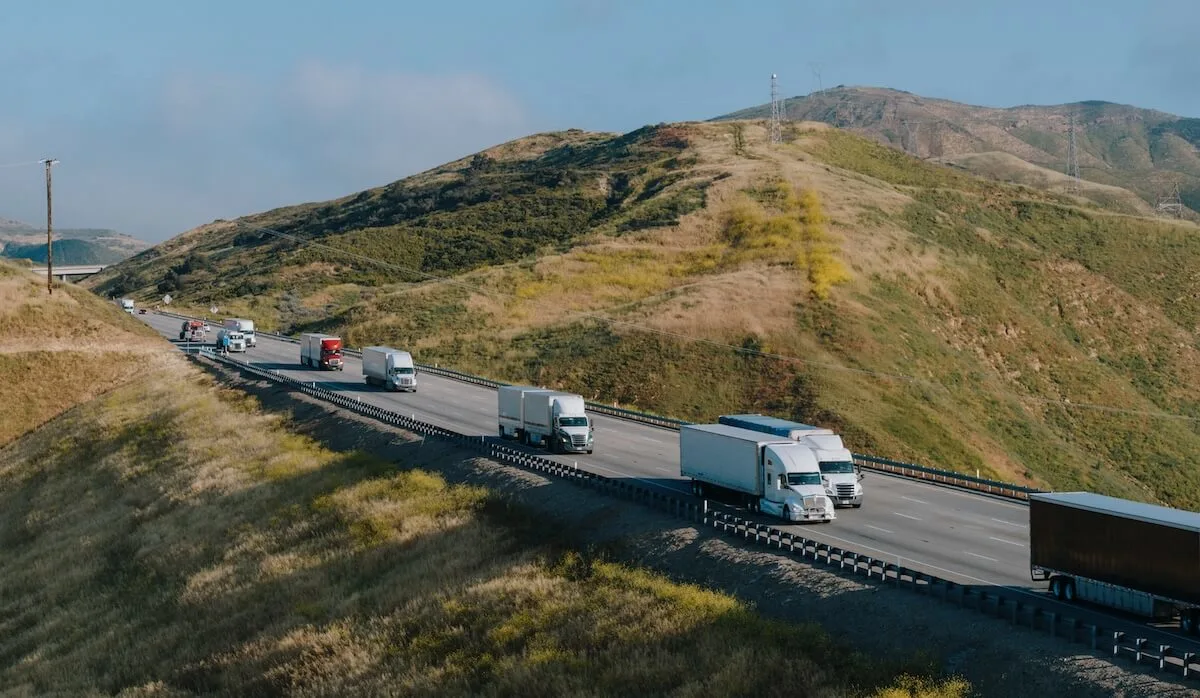 Semi-remorques circulant sur une autoroute panoramique à plusieurs voies longeant des collines verdoyantes, représentant le transport de fret actif.