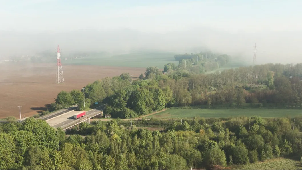 Aerial view of a highway bridge with red truck crossing through foggy countryside with farmland and transmission towers.
