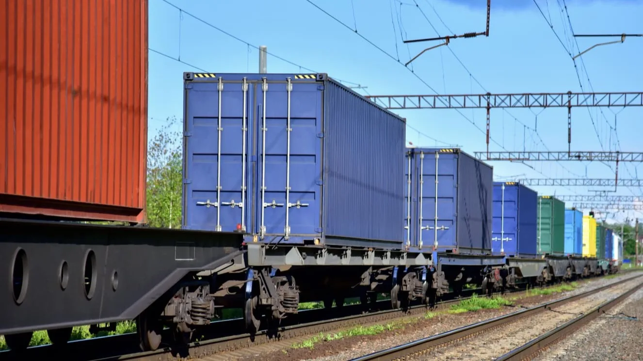 Freight train carrying colorful shipping containers on flatbed cars, with blue containers prominent under overhead power lines.