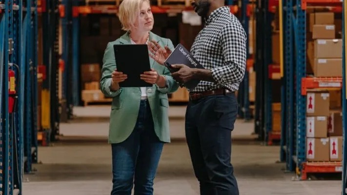 Two professionals discussing inventory in a warehouse with blue shelving racks and stacked cardboard boxes.