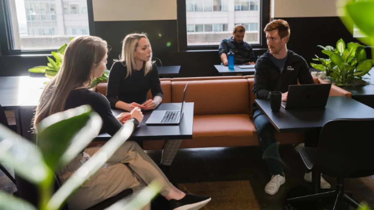 Four people working in a modern office space with laptops, orange seating, large windows, and green plants.